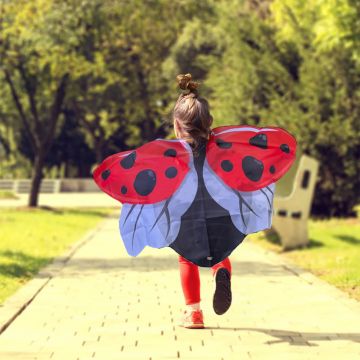 A child running down a path surrounded by trees, wearing a red ladybird wings cape with black spots